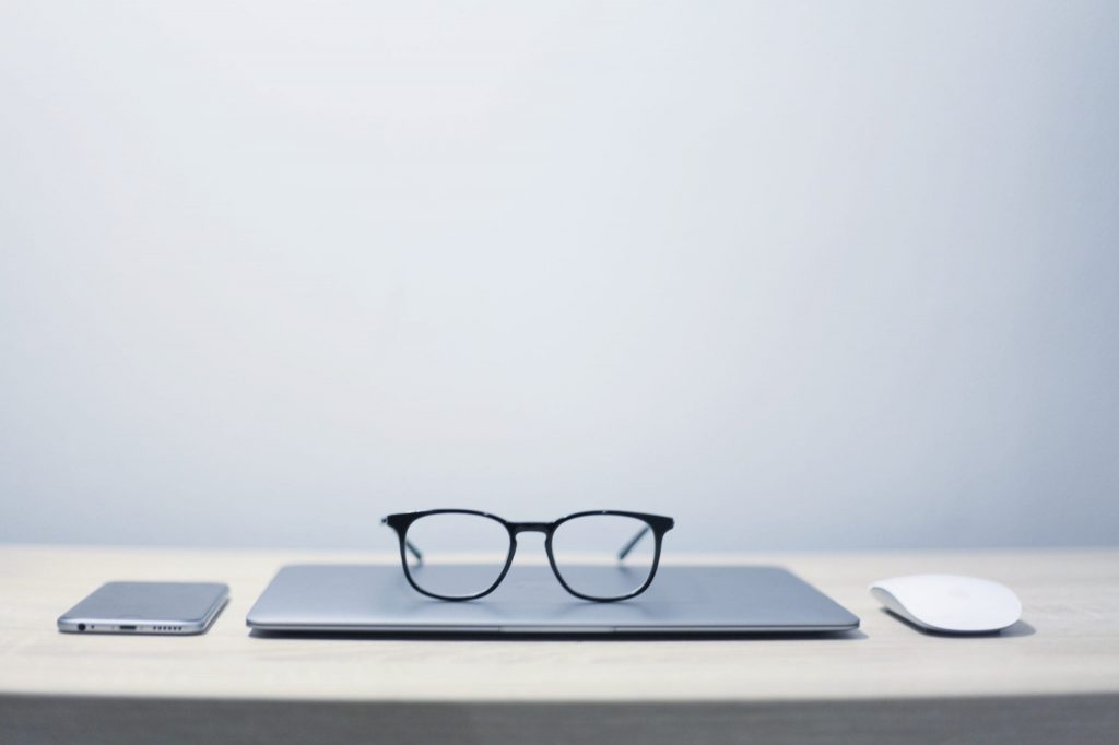 a shot of a glasses on a silver laptop with a mouse on the right and a phone on the left and all rests on a table