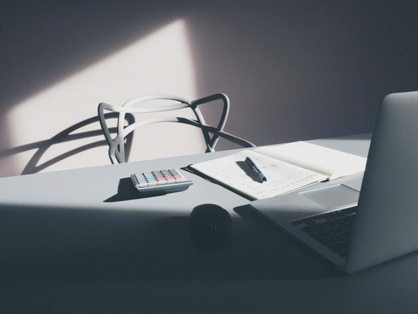 a photo of a workstation with a chair behind a table and a laptop, a notebook, a pen, a calculator, and a mouse on the table with a ray of light shines upon the chair