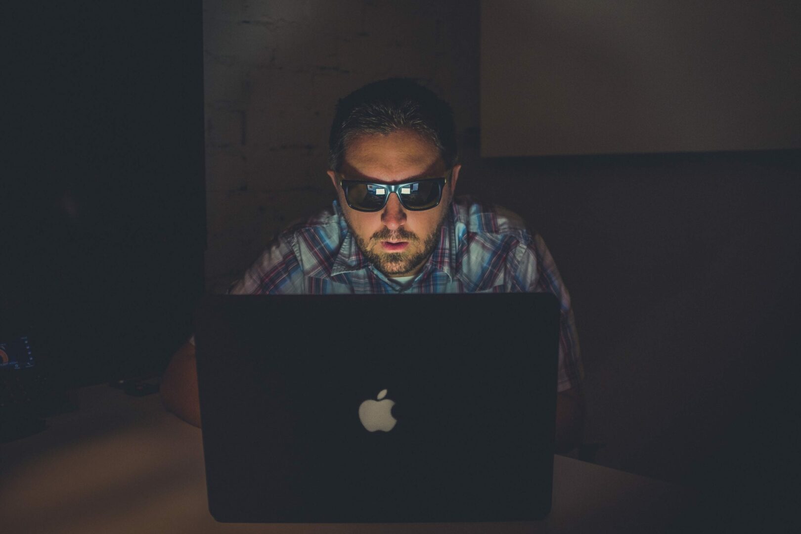 a shot of a man sitting in front of his laptop watching the laptop in the dark with sunglasses on