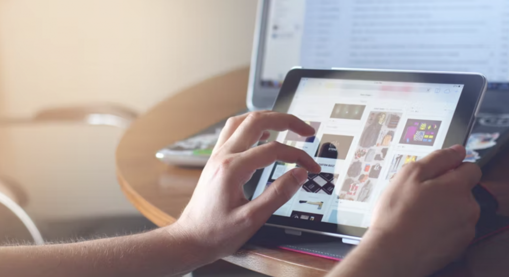 a shot of a person holding a tablet and using it in front of a laptop on a table