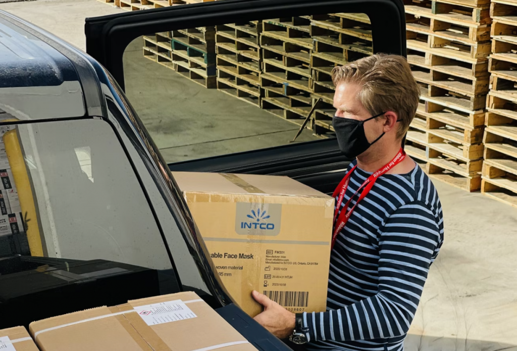 a shot of a man wearing striped black and white shirt with a red lanyard carrying a box into a black car