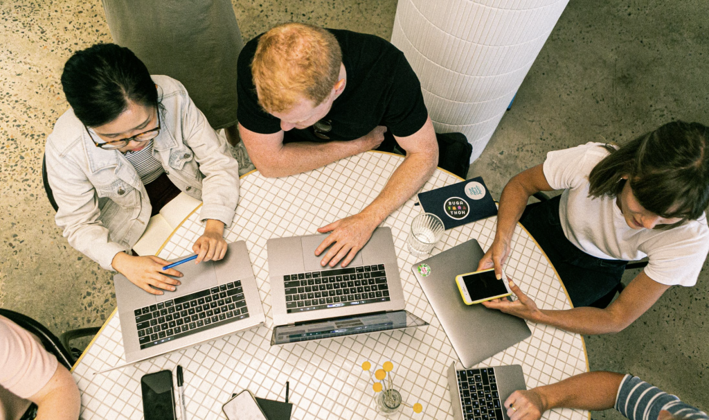 an aerial shot of a few people on a circular table with laptops on the table discussing with each other