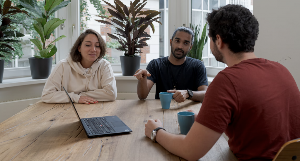 a shot of 2 men and a woman discussing over a table with a laptop on the table and each man has a mug with them