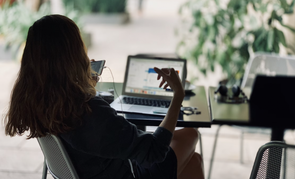 a shot of a woman talking to a phone with her left hand sitting on a chair in front of her laptop on a table.