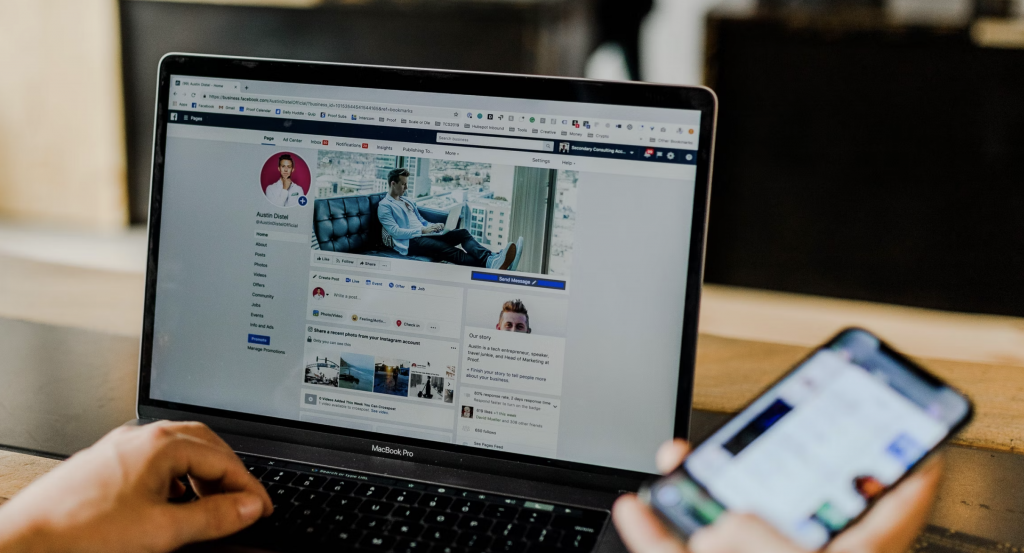 a shot of a person using a laptop with left hand and the phone on the right hand with the screen showing a facebook profile of a man named Austin Dishel