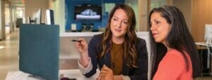 a photo of 2 women sitting side by side while looking at a monitor in an office
