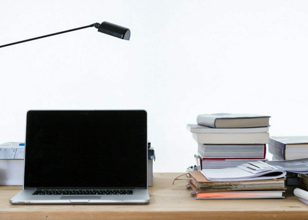 an image of a laptop on the left on a table and a stack of books on the right with a lamp hanging above the laptop