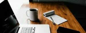 a shot of a laptop, with a mug of coffee and a notebook and a pen on top of a wooden table.