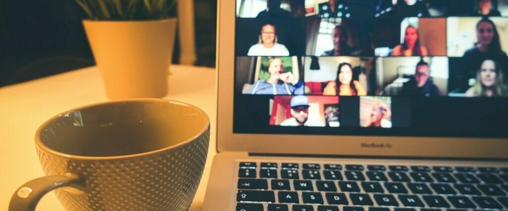 an image of a macbook air with the screen showing a few people on a video conference and a mug with handle besides the macbook