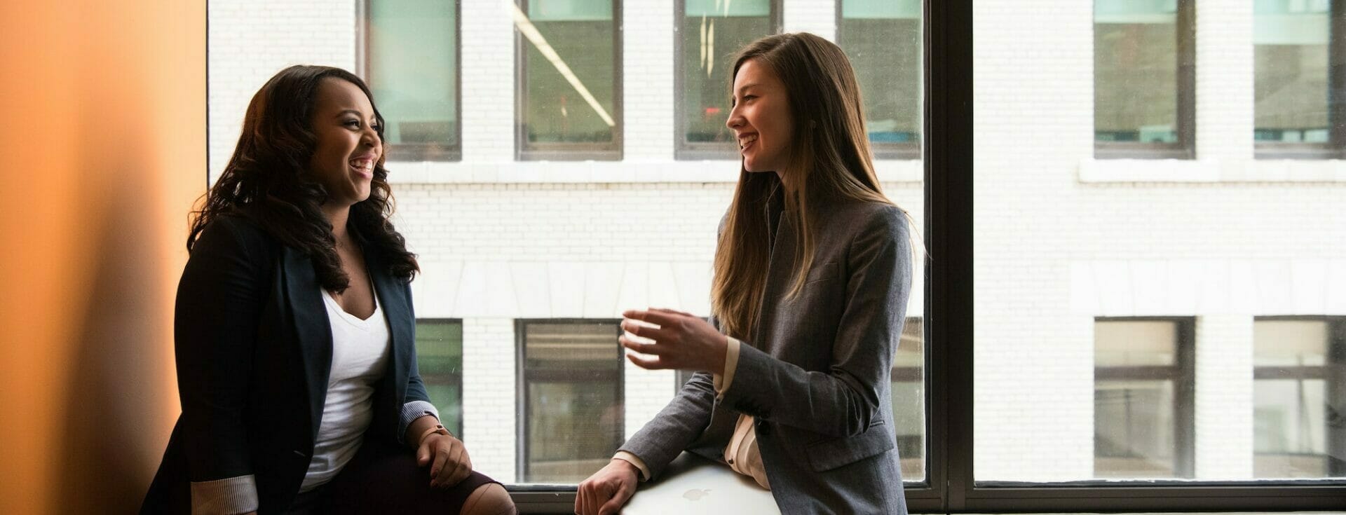 two women sitting on the ledge by the window talking and smiling at each other in a brightly lit space