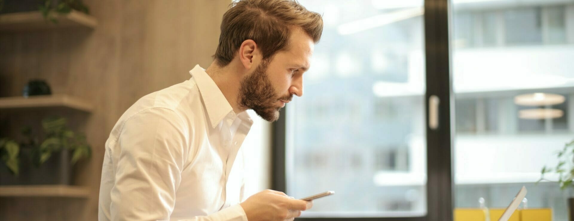 a shot of a man holding a phone looking at a laptop in a brightly lit room