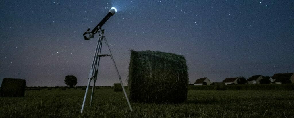 a shot of a telescope in a field with a haystack behind it at night with the sky full of stars